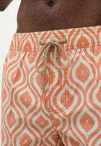 Un maillot de bain orange et crème avec un motif en ondulation comportant de petits crânes. Taille élastique avec un cordon de serrage tressé à nouer, texture lisse.