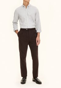 Camicia button-up grigia chiara abbinata a pantaloni pieghettati marrone scuro e mocassini neri. Il tessuto sembra liscio con una vestibilità strutturata.