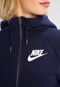 Námořnická mikina na zip s jemnou texturou, s kapucí na šňůrku a bílým logem Nike na hrudi. Obsahuje kovový zip a boční kapsy.