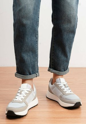Personne portant un jean bleu foncé retroussé et des baskets blanc-gris, debout sur un sol en bois.