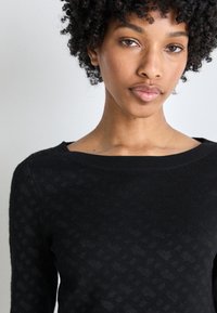 Pulover negru tricotat, cu un model texturat, guler larg și mâneci lungi. Țesătura pare moale și ușor elastică.