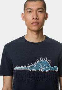 Námořnické bavlněné tričko s modrou grafikou dinosaura a textem "SLEEPSAURUS" v texturovaných písmenech na přední straně. Krátké rukávy, kulatý výstřih.