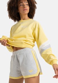 Sudadera amarilla recortada con logotipo bordado y detalles a rayas grises, combinada con pantalones cortos grises con rayas laterales amarillas y logotipo.