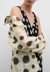 Robe noire sans manches avec des pois blancs, superposée d'un châle en dentelle crème avec de grands pois noirs.