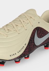 Μπεζ ποδοσφαιρικό παπούτσι Nike με μαύρο και κόκκινο σχέδιο λεοπάρδαλης στη φτέρνα, ασημένιο λογότυπο Nike swoosh, μπεζ κορδόνια και μαύρα καρφιά με κόκκινες άκρες.