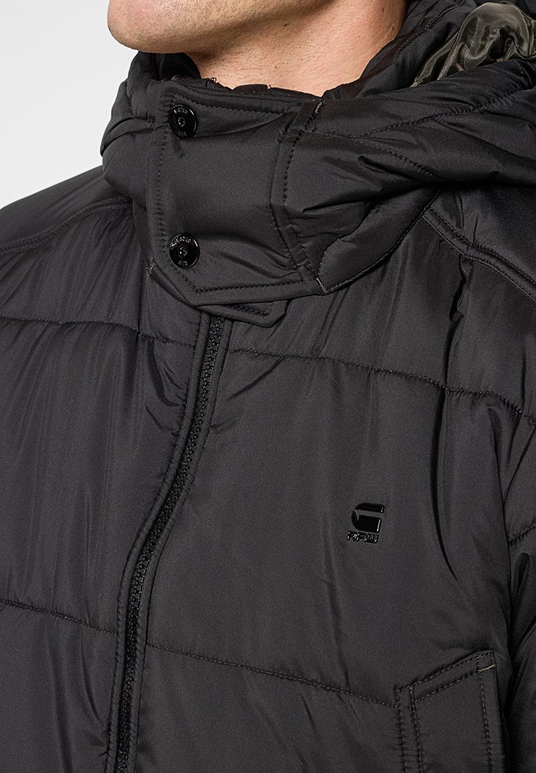 Chaqueta acolchada negra con un alto cuello, cierre de botones, cremallera frontal y un pequeño detalle de logo. Textura suave y diseño aislante.