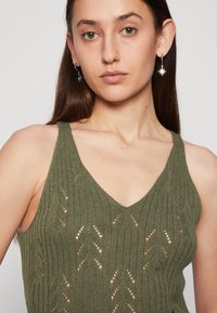 Débardeur en maille vert olive avec un décolleté en V, des motifs en chevrons et des détails en point ouvert. Le mannequin porte des boucles d'oreilles en forme d'étoile argentée.