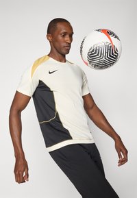Jersey de Nike texturizado en blanco y negro con acentos dorados, de mangas cortas y forma ajustada; jugador haciendo malabares con un balón de fútbol con patrón.