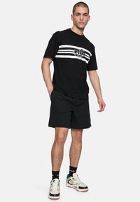 Schwarzes Kurzarmhemd mit weißen horizontalen Streifen, ausgestattet mit einem Logo und schwarzen Shorts. Kombiniert mit gemusterten Turnschuhen und Sneakersocken.