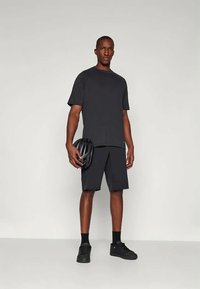 Άνδρας φορεί μαύρη μπλούζα, μαύρα σορτς, μαύρα αθλητικά παπούτσια και κάλτσες, κρατώντας ένα μαύρο κράνος ποδηλασίας με φόντο απλής υφής.