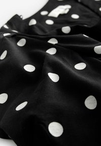 Tessuto nero con grandi pois bianchi, texture liscia e drappeggio morbido. Include un'etichetta nell'angolo in alto a sinistra.