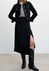 Giacca in denim nera sopra una camicia a righe bianco e nere, abbinata a una gonna nera con uno spacco laterale e stivali da cowboy neri con tacco.