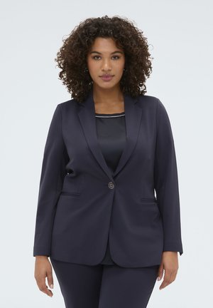 Vrouw met krullend haar, die een donkerblauwe blazer draagt over een zwarte top, staande tegen een effen lichtgrijze achtergrond.