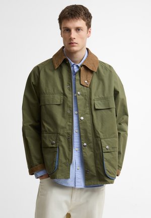 Noor mees, kes kannab oliivrohelise jope pruunist sametkraega, sinise nööpidega särgi ja valgete pükstega, seisab valge tausta ees.