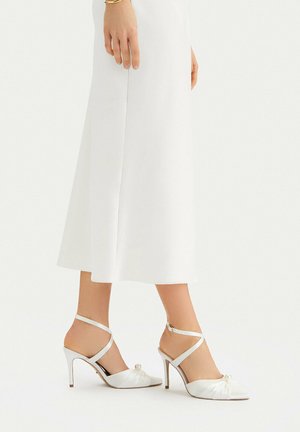 Mujer con falda midi blanca y zapatos blancos de tacón alto con tiras en el tobillo y detalle de perlas en los dedos.