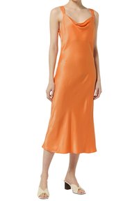 Pomarańczowa satynowa sukienka na ramiączkach z dekoltem typu cowl, cienkimi ramiączkami i płynna sylwetką, sięgająca do połowy łydki. Modelka nosi beżowe mule na słupku.