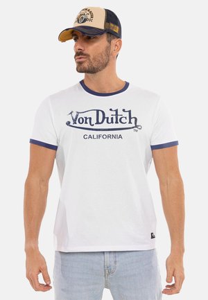 Homme portant un t-shirt blanc Von Dutch California avec une bordure bleue et une casquette trucker beige et bleu marine, regardant sur le côté.