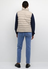 Beige gewatteerd vest met hoge kraag en verticale stiksels, gedragen over een navyblauw shirt met lange mouwen en gecombineerd met lichtblauwe spijkerbroek en zwarte schoenen.