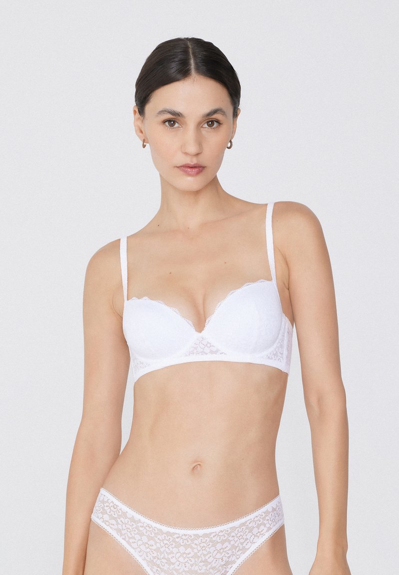 Soutien-gorge en dentelle blanche avec des bords festonnés et des fines bretelles, présentant un décolleté plongeant et un motif floral, assorti à un slip en dentelle.