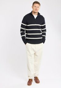 Pull en maille navy avec rayures blanches, col zippé, associé à un pantalon crème et des chaussures marron. La texture est côtelée et tissée.
