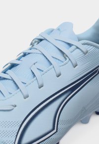 Chaussure de football bleu clair avec des lacets blancs et un logo Puma bleu foncé sur le côté, montrant une partie de la semelle à crampons.
