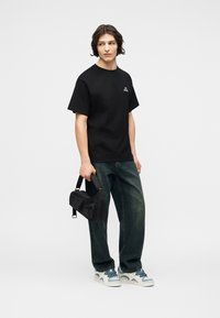 Νεαρός άνδρας με μαύρο μπλουζάκι, φαρδιά σκούρα τζιν, λευκά και μπλε αθλητικά παπούτσια, κρατώντας μια μαύρη τσάντα χιαστί μπροστά σε απλό φόντο.