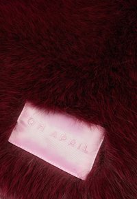 Dirbtinis kailis sodriai burgundiškoje spalvoje, turintis ilgas, minkštas skaidulas. Šone matoma rožinės spalvos etiketė su įspaustu tekstu "OH APRIL".