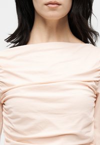 Γυναίκα με μεσαίου μήκους σκουρόχρωμα μαλλιά που φοράει ροζ ανοιχτή στη μέση ρουχισμένη μπλούζα, εμφανίζοντας το λαιμό και το πάνω μέρος του κορμού.