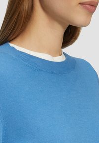 Blauer leichter Pullover mit rundem Halsauschnitt; verfügt über eine feine Stricktextur und gerippte Ärmelsäume, über einem weißen Hemd getragen.