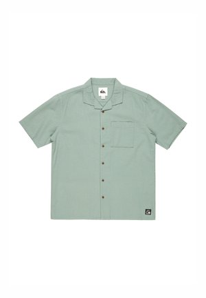 Short-sleeve button-up shirt in light green textured fabric, featuring a chest pocket and brown buttons. Logo patch at the bottom left.