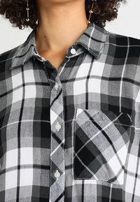 Black and white plaid button-up shirt featuring a classic collar, chest pocket, and white buttons on a textured fabric.
