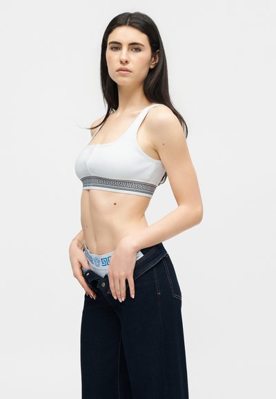 Νέα γυναίκα με μακριά σκούρα μαλλιά ρυθμίζει σκούρο τζιν με ξεκούμπωτο κουμπί, φοράει λευκό κοντό τοπ και ζώνη με σχέδιο ελληνικού κλειδιού.