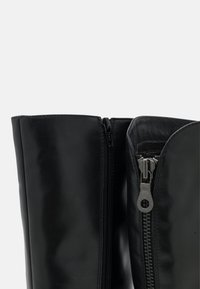 Bottes en cuir noir jusqu'aux genoux avec une texture lisse ; dotées de double zippers à l'avant pour faciliter l'enfilage. Design élégant et minimaliste.