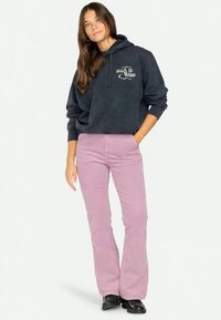 Felpa blu navy con logo bianco sul petto, abbinata a pantaloni in velluto a coste rosa chiaro a campana e calzature nere. Outfit semplice e casual.