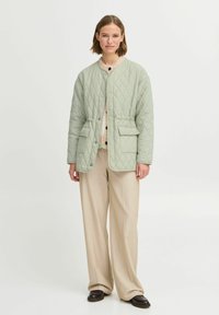 Chaqueta acolchada de color verde claro con cuello redondo, bolsillos frontales y cierres a presión, llevada encima de un suéter texturizado y pantalones de pierna ancha color beige.