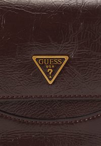 Portafoglio sintetico marrone con texture, caratterizzato da un logo triangolare dorato di Guess USA con un punto interrogativo, dettagli cuciti e bordi lisci.