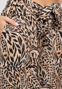 Tissu imprimé animal dans des nuances de beige, noir et marron. Présente un détail de nœud papillon à la taille avec des poches intégrées dans le design.