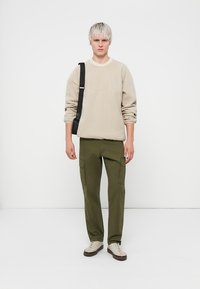 Sweatshirt beige avec col rond, pantalon cargo olive avec poches latérales, et baskets beiges. Le modèle porte un sac noir en bandoulière.