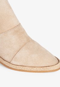Bota de ante beige con punta afilada, que presenta correas cruzadas y una suela trenzada con yute para añadir textura y detalle.
