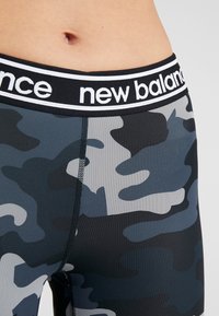 Detailní záběr na pas osoby, která má na sobě černé a šedé maskáčové legíny s černým pasem, na kterém je bílé logo "new balance".