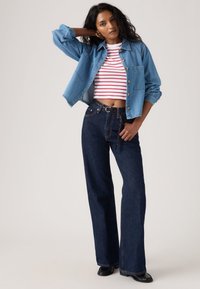 Naine seisab käsi pea taga, kandes teksajopet, punaseid ja valgeid triibulisi kärbitud topi, tumedaid pükse ja musti kingi ühevärvilise taustaga.