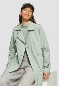 Trench coat de doble botonadura en color verde claro con botones grandes. Llevado sobre una blusa gris texturizada y pantalones grises con estampado, complementado con una gorra gris.