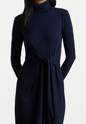 Femme portant une robe à manches longues et col roulé bleu marine avec un détail noué à la taille.