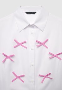 White collared shirt featuring six pink ribbon accents. Smooth fabric with button closure, designed for a tailored fit and clean lines.