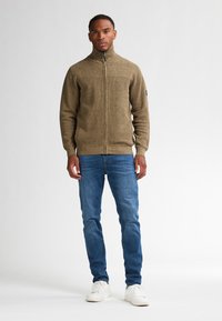 Sudadera de punto marrón con cremallera y cuello alto, combinada con vaqueros azules y zapatillas blancas. Presenta una textura sutil y un diseño ajustado.