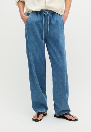 Personne portant un pantalon large en denim bleu avec taille élastiquée et sandales noires à brides, mains dans les poches, se tenant devant un fond uni.