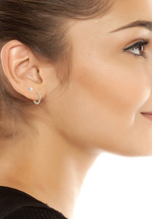 Nuoren naisen oikea profiili, jossa helmirouva- ja pienet timanttikorvakorut, sileä iho, tumma tukka vedetty taakse, lievä hymy.