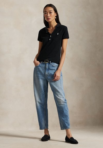 Žena stojící s jednou rukou v kapse, oblečená v černé polokošili, světle modrých džínách, černém opasku, černých mokasínách a zlatých náušnicích.