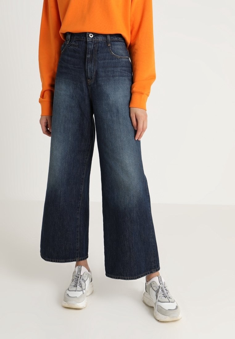 Szerokie ciemnoniebieskie dżinsy o wysokim stanie, zestawione z jaskrawopomarańczowym swetrem i szarymi sneakersami. Gładka faktura z minimalnym blaknięciem.