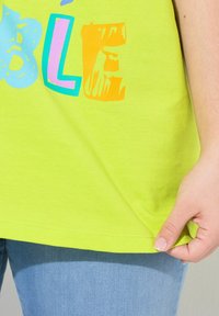 Limonkově zelené tričko s barevným textem "ABLE", s měkkou bavlněnou texturou a volným střihem, částečně zobrazené s rukou, která drží lem.